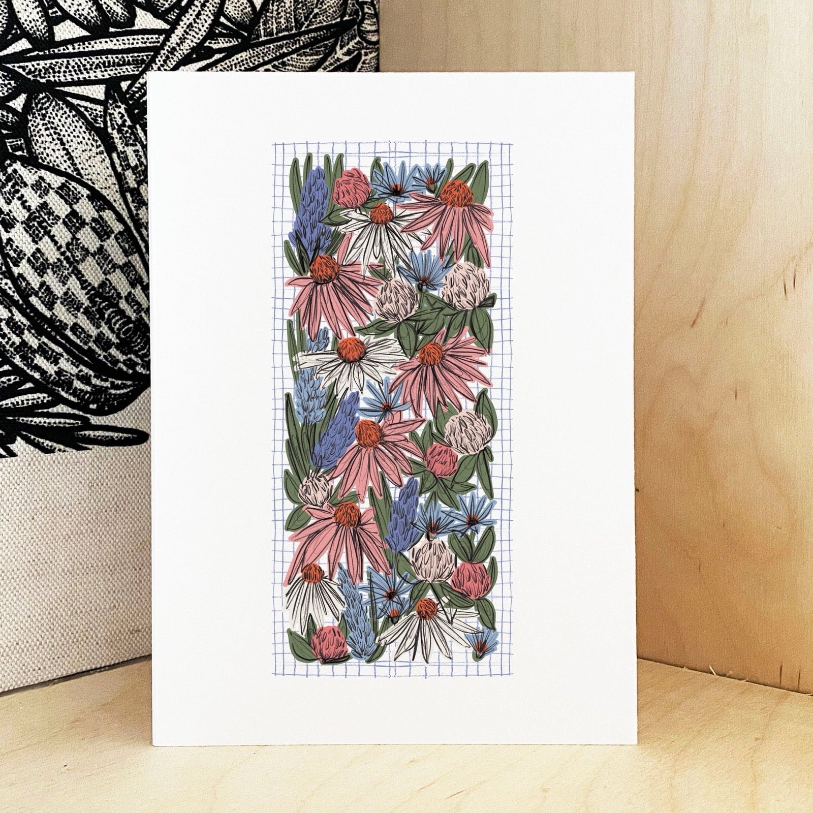 Floral artwork on a grid background with a wooden surface and patterned fabric in the background.