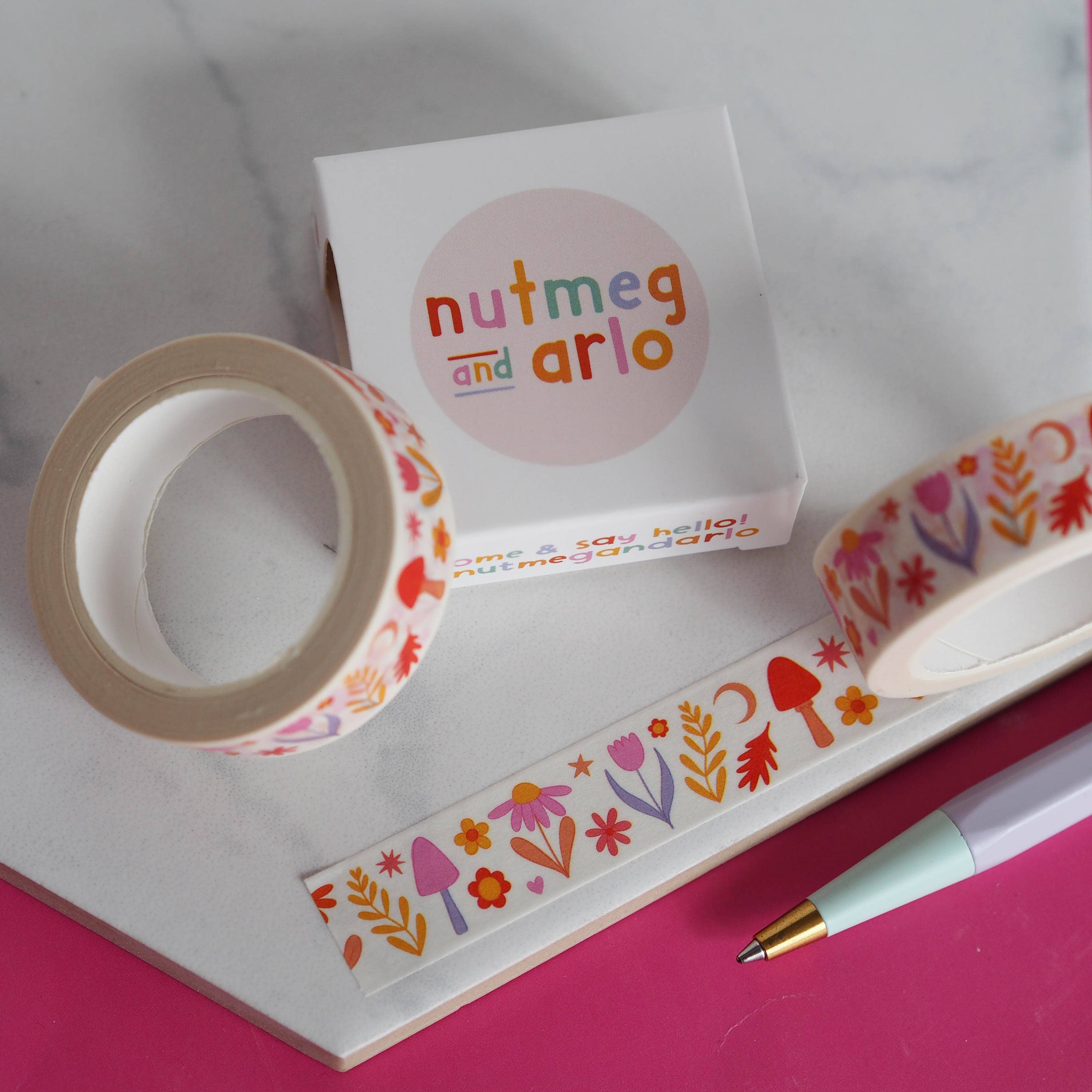 Decorative tape with floral designs and a box labeled 'nutmeg and arlo' on a pink surface.