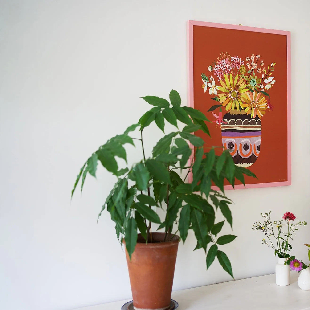 Potted plant on a white surface with a colorful floral painting in a pink frame on the wall.