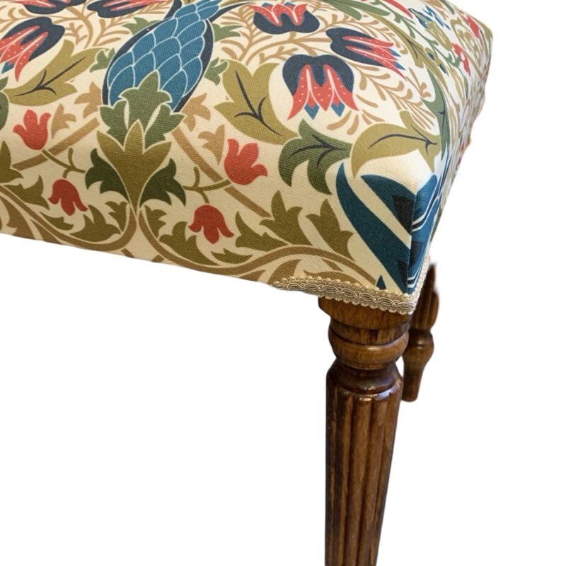 A rectangular bench covered in cream linen fabric with a multicoloured floral William Morris style design, featuring mahogany legs and cream binding.