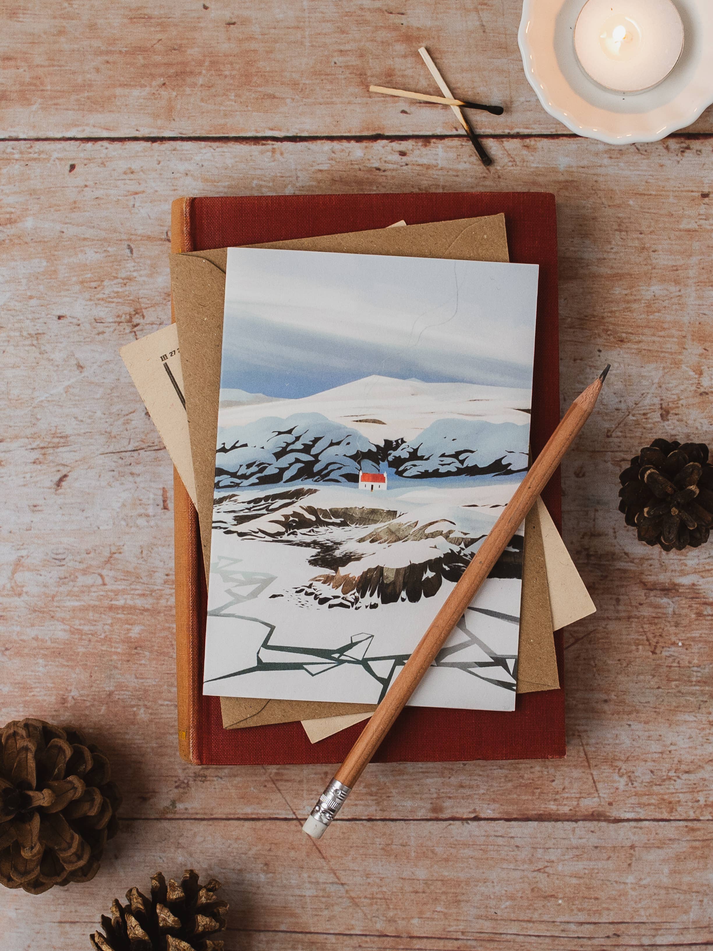A greeting depicting a snowy mountain landscape with a small red house, on a rustic wooden table with a red book, kraft envelopes, a pencil, pinecones, and a lit candle.