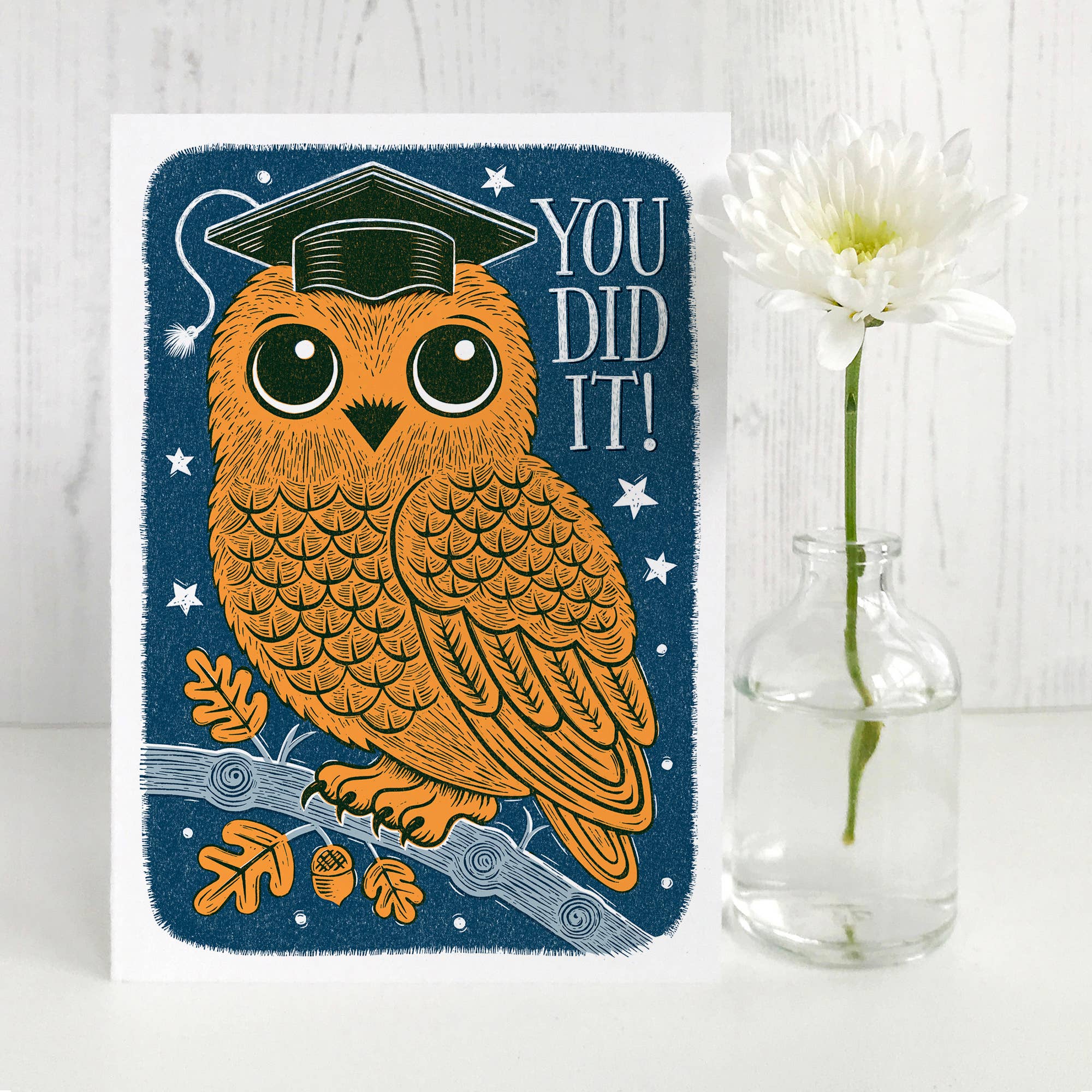 Card with an owl wearing a graduation cap and 'YOU DID IT!' text, next to a vase with a flower on a light wooden surface.
