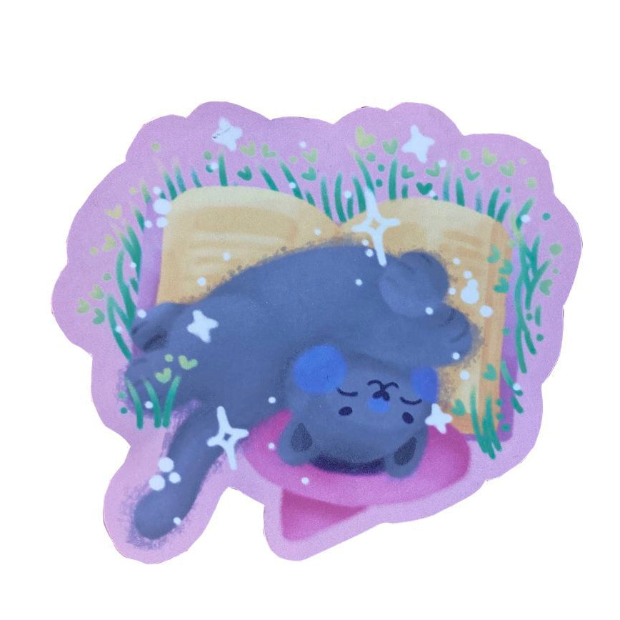Handmade sticker of a grey witch cat with a pink witch's hat, sleeping on an open book.