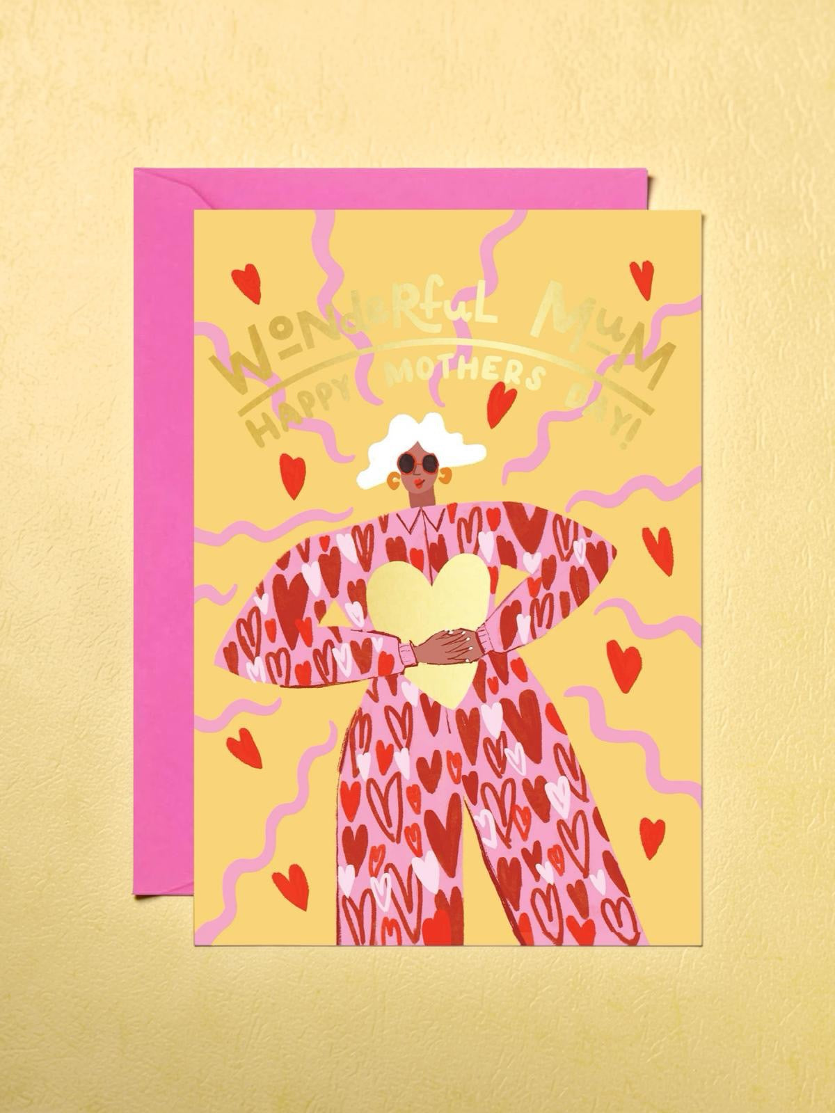 Mother's Day card featuring an illustration of a fabulous mum with white hair, wearing a red jumpsuit covered in love hearts, holding a giant gold love heart, on a yellow background.