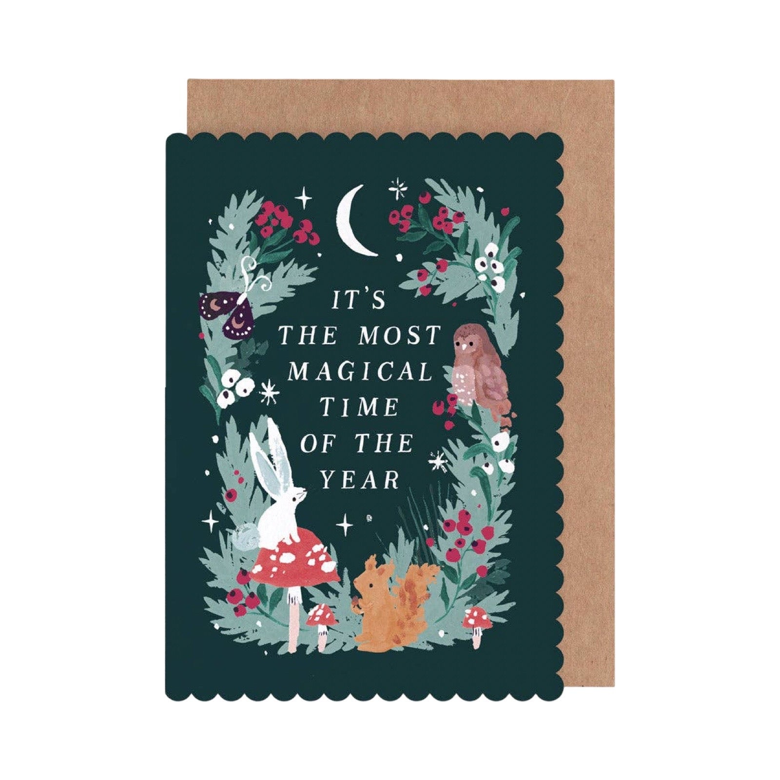 A dark teal greeting card with scalloped edges featuring a whimsical woodland illustration of a rabbit, owl, fox, mushrooms, and stars, with the text 'IT'S THE MOST MAGICAL TIME OF THE YEAR'. A kraft paper envelope is partially visible behind it on a white background.