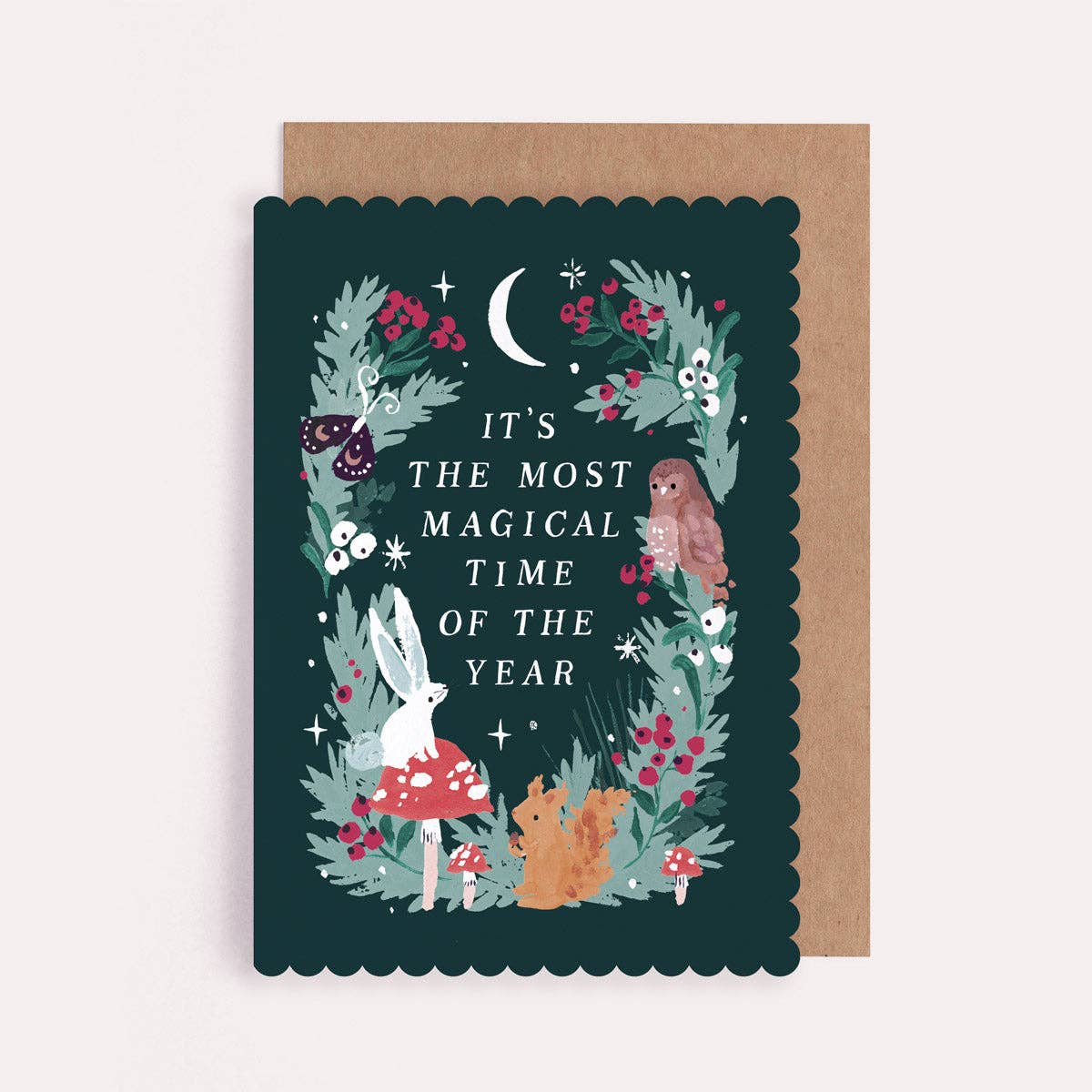A decorative greeting card with a dark teal background and scalloped edges, featuring whimsical woodland illustrations including a white rabbit in a red mushroom cap, a brown owl, a fox, berries, stars, and the text 'IT'S THE MOST MAGICAL TIME OF THE YEAR.' A plain kraft envelope is placed behind the card on a light neutral background.