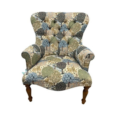 A cream linen fabric armchair with a pattern featuring brown deer and trees in shades of light blue, navy, and green, button-back design, with mahogany legs and gold castors.