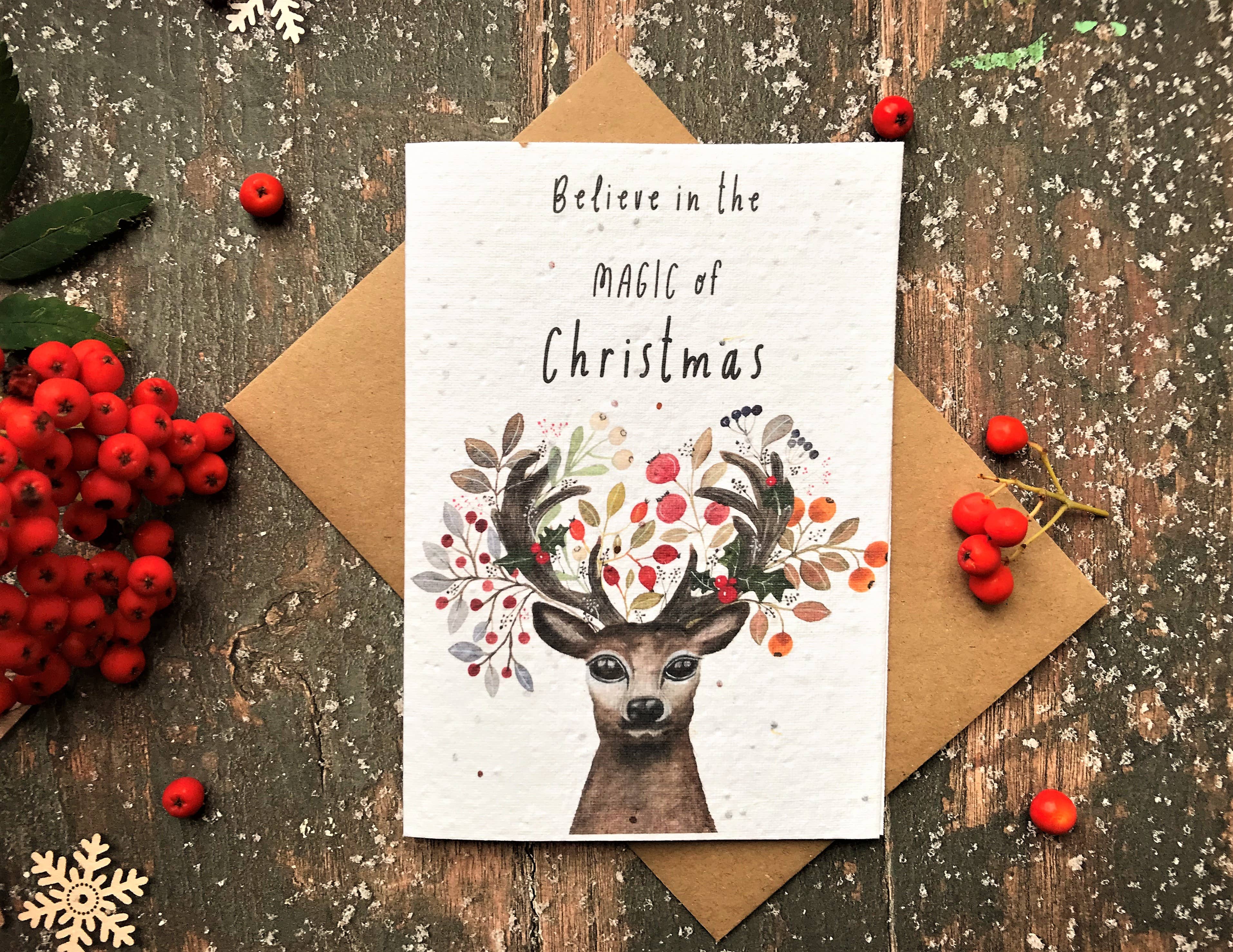 A Christmas greeting card with a watercolour deer and antlers made of leaves and berries, reading 'Believe in the Magic of Christmas,' styled on a kraft envelope over a rustic wooden surface with red berries and snowflake decorations.