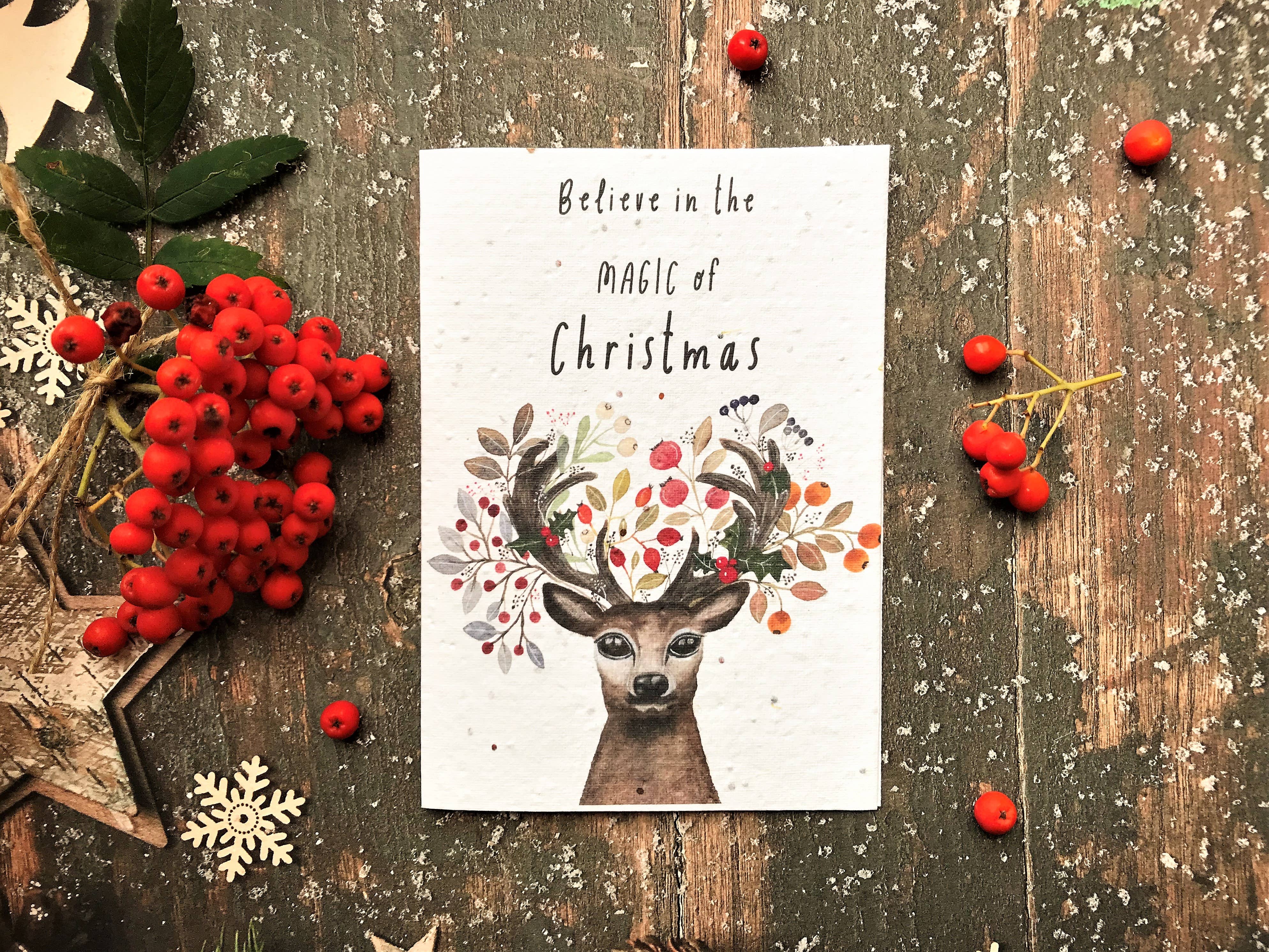 A white Christmas card featuring a deer with antlers decorated with berries and leaves, and the text 'Believe in the magic of Christmas'. The print is styled in a flat-lay on a rustic wooden surface surrounded by red berries, green leaves, and snowflake decorations.