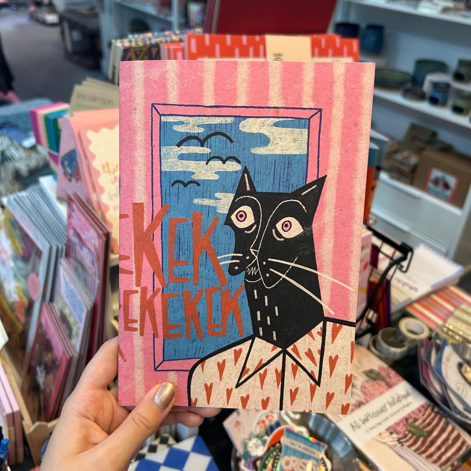 A hand holds a pink and orange illustrated card featuring a stylized black cat and the text 'Kek kekok' against a store background filled with colourful items.