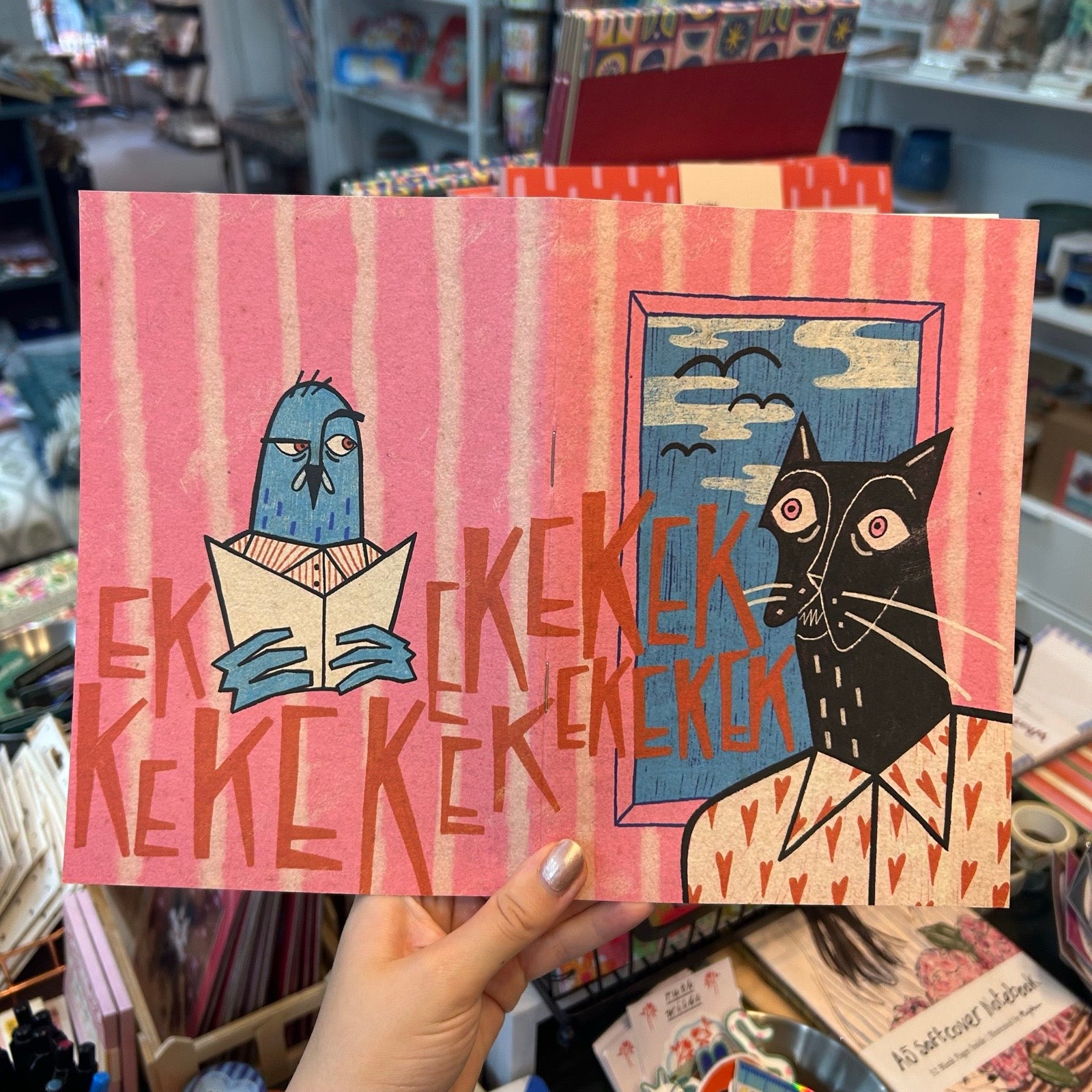 A hand holds a pink illustrated art print featuring a blue bird reading and a black cat looking out a window, with stylized text across the centre, in a retail store setting.