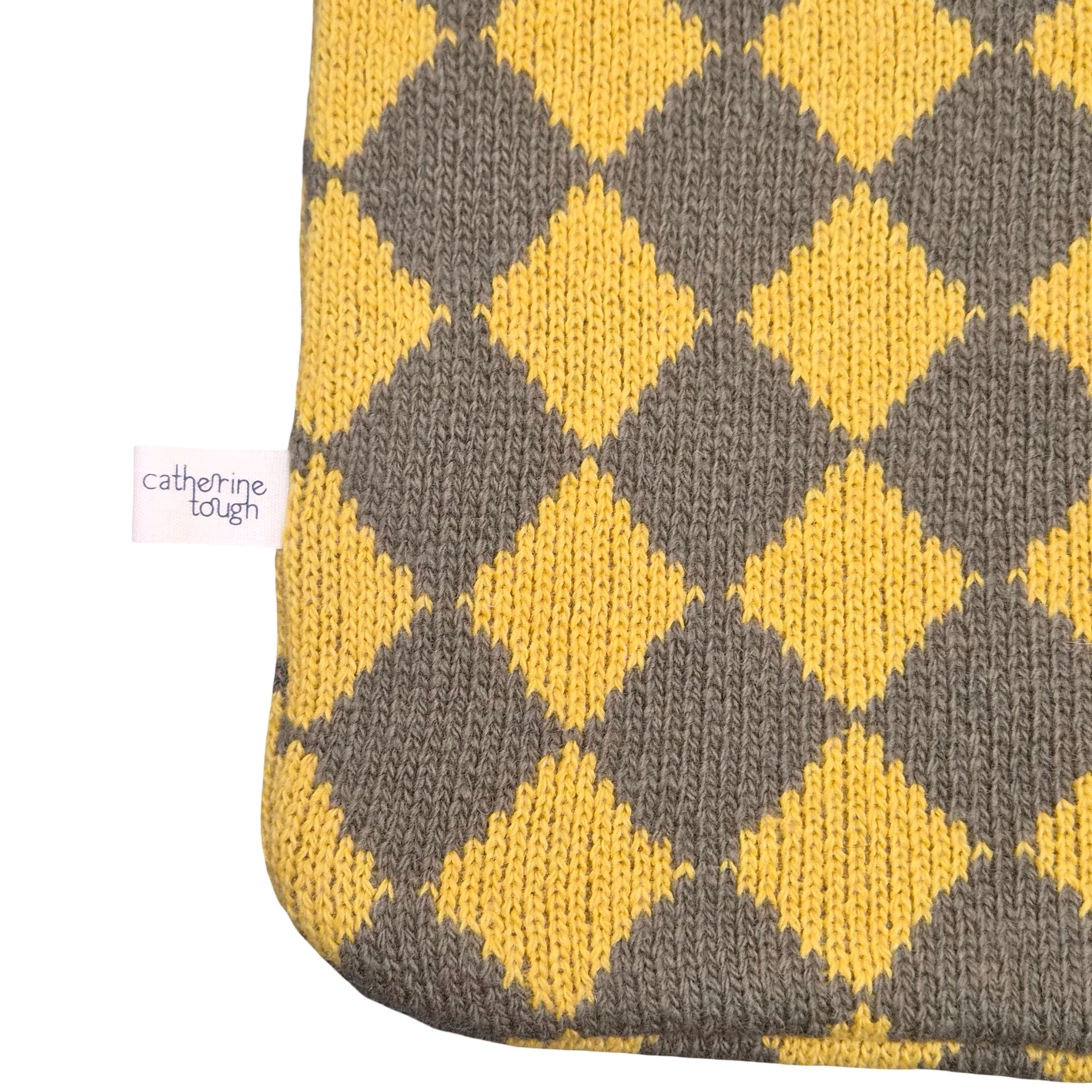 Close-up of a knitted hot water cover in yellow and grey argyle pattern, showing a small white tag that reads 'catherine tough' on a white background.