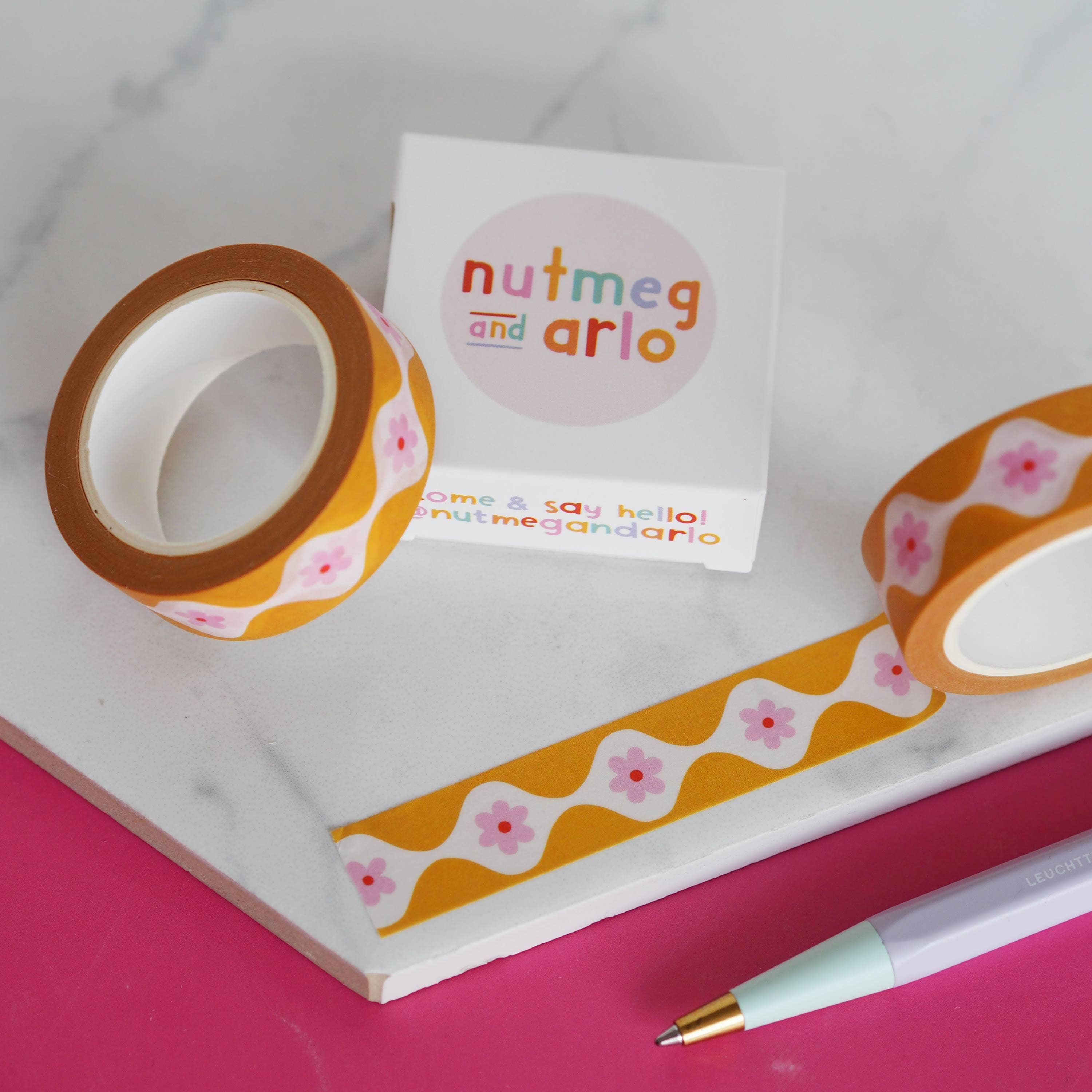 Decorative tape, pen, and notepad with 'nutmeg and arlo' branding on a pink surface.