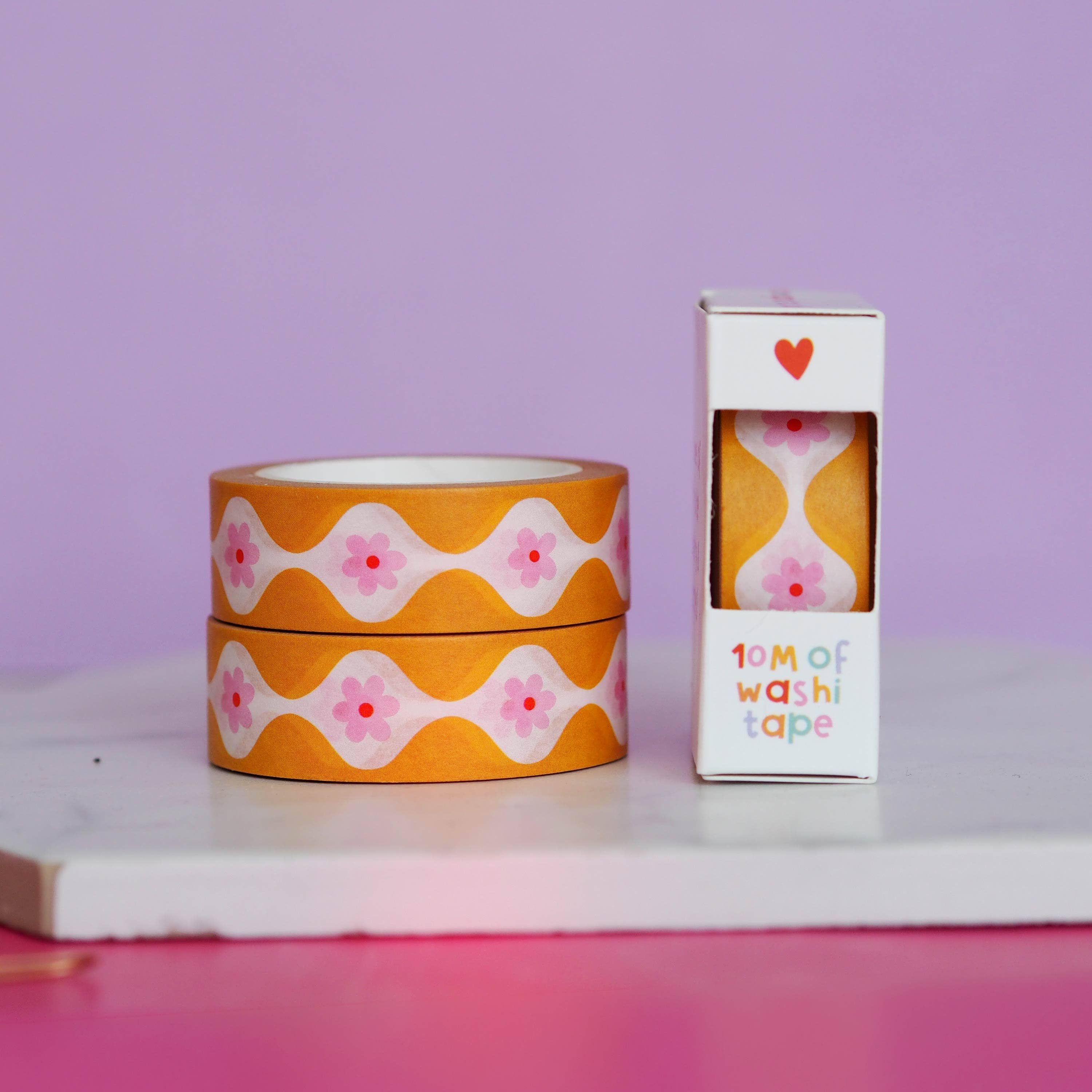 Two rolls of orange washi tape with pink floral patterns and a box labeled '10m of washi tape' on a purple background.